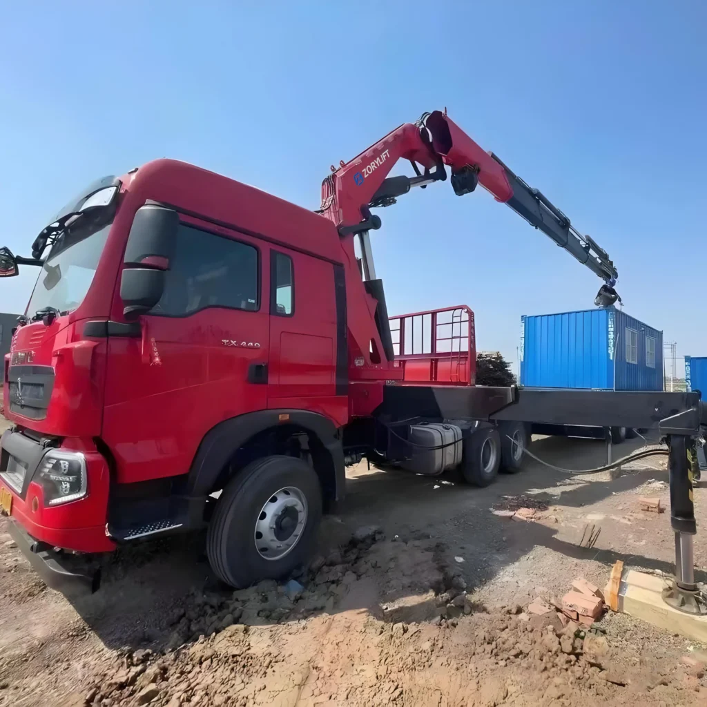 Close-up of a red heavy-duty truck equipped with an articulated boom crane (knuckle boom crane), a type of truck mounted crane, being used by a customer for a lifting task involving a blue module on a worksite.