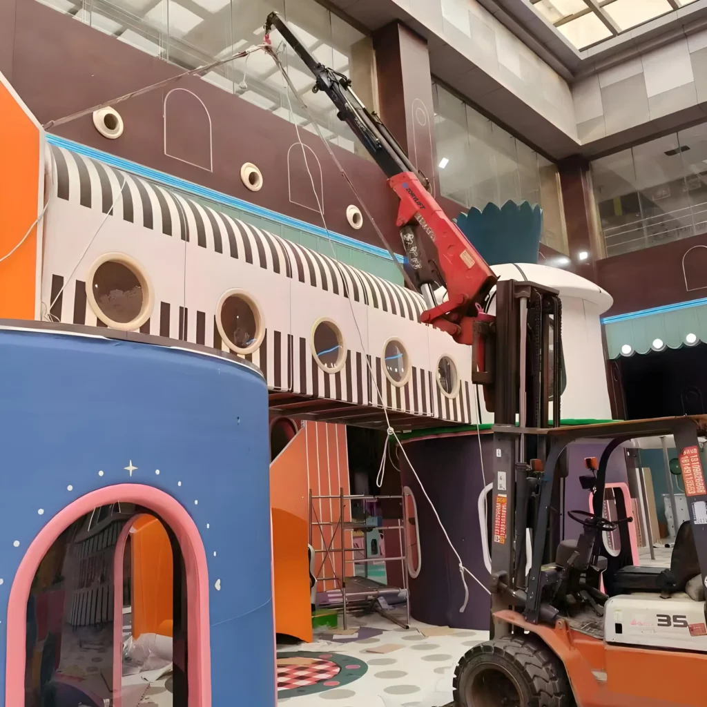 A forklift crane, a type of specialty crane, assisting a customer with a heavy lift during the installation of a large, colorful structure indoors. This demonstrates a common application in construction or facility setup.