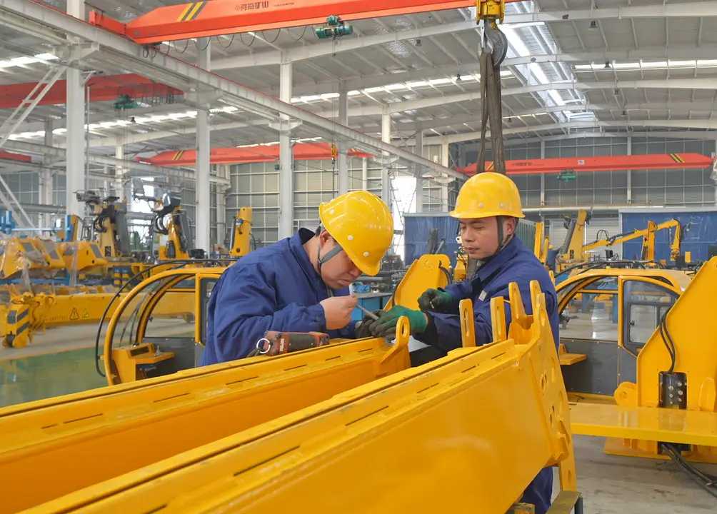 Inside our modern factory workshop, an experienced team of technicians is meticulously assembling the main structure of a large yellow vehicle mounted crane, showcasing our strong manufacturing capabilities.